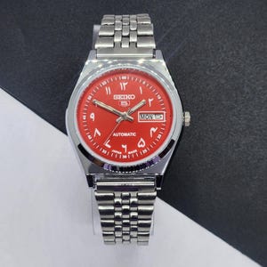 Könnte beinhalten: Eine silberfarbene Seiko 5 Automatik-Armbanduhr mit rotem Zifferblatt. Die Uhr verfügt über arabische Ziffern, eine Tages- und Datumsanzeige und ein Edelstahlarmband. Das Zifferblatt zeigt den Tag als "MON 5".