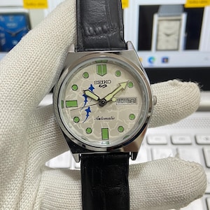 Puede incluir: Un reloj de pulsera automático Seiko plateado con una esfera blanca y una correa de cuero negra. La esfera tiene un diseño verde y azul con las palabras "Seiko Automatic" y "MON 18".