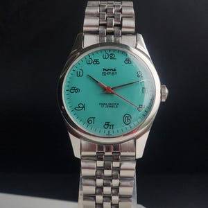 May include: A silver-toned wristwatch with a teal face and a matching silver band. The watch face features black text and numerals, with red second hand. The watch is labeled "PARA SHOCK 17 JEWELS".