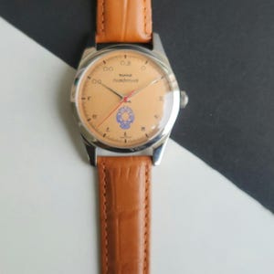 K&ouml;nnte beinhalten: Eine Vintage-Armbanduhr mit hellorangefarbenem Zifferblatt, silbernem Geh&auml;use und braunem Lederarmband. Das Zifferblatt der Uhr hat schwarze Ziffern und ein blaues Emblem. Das Armband ist hellbraun mit Krokodilmuster.