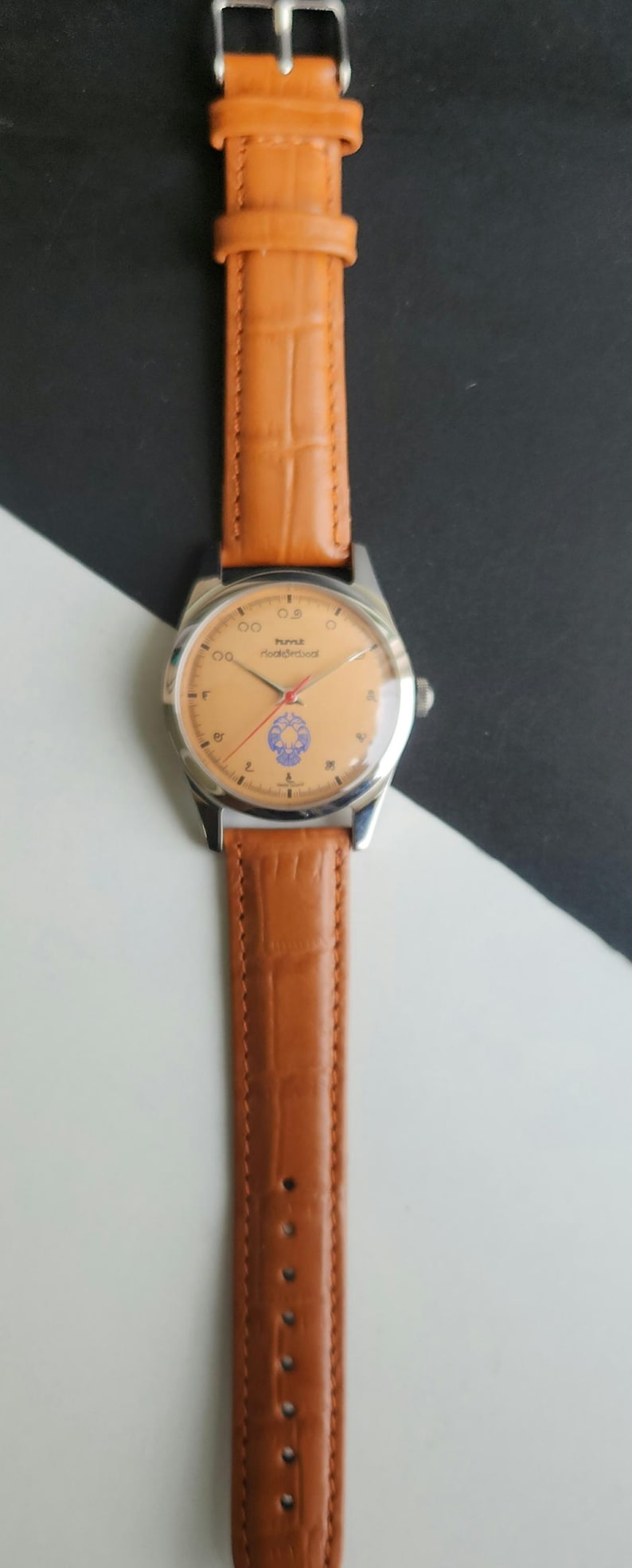 K&ouml;nnte beinhalten: Eine Vintage-Armbanduhr mit hellorangefarbenem Zifferblatt, silbernem Geh&auml;use und braunem Lederarmband. Das Zifferblatt der Uhr hat schwarze Ziffern und ein blaues Emblem. Das Armband ist hellbraun mit Krokodilmuster.