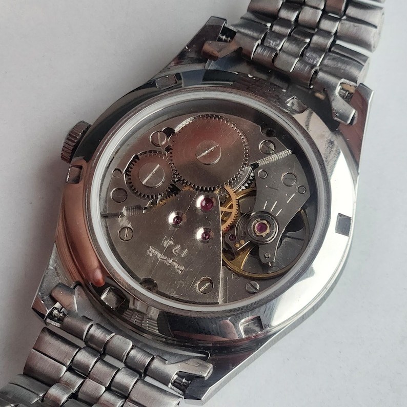 May include: A silver-toned wristwatch with a visible mechanical movement. The watch's back reveals gears, jewels, and intricate internal components. The watchband is made of interlocking metal links. The watch is a vintage style.