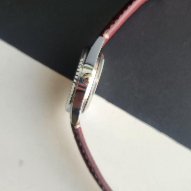 May include: A silver wristwatch with a brown leather band. The watch face is visible, and the bezel has a textured edge. The band is a deep reddish-brown color. The watch is angled, showing the side profile against a black and white background.