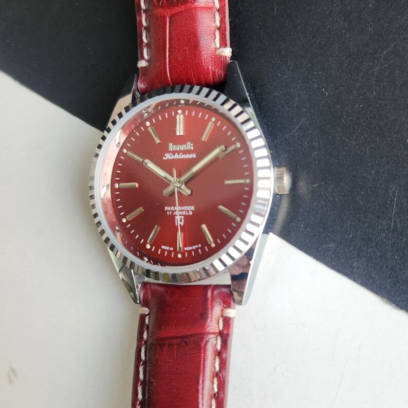 May include: A vintage wristwatch with a burgundy face and a silver bezel. The watch has a red leather band with white stitching. The watch face displays the brand name and the words "Parashock 17 Jewels."