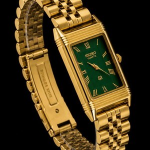 May include: A gold-tone rectangular wristwatch with a green dial, featuring gold Roman numeral hour markers and gold hands. The watch has a multi-link metal bracelet with a clasp engraved with the words STAINLESS STEEL. The Seiko Quartz logo is visible.