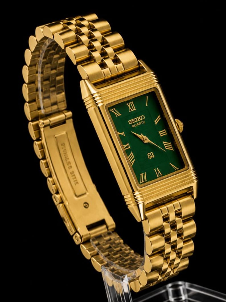 May include: A gold-tone rectangular wristwatch with a green dial, featuring gold Roman numeral hour markers and gold hands. The watch has a multi-link metal bracelet with a clasp engraved with the words STAINLESS STEEL. The Seiko Quartz logo is visible.