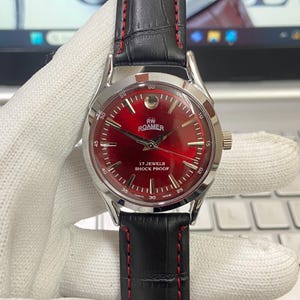May include: A vintage RW Roamer watch with a bright red face and silver casing. The watch has a black leather strap with red stitching. The face displays the text "17 Jewels Shock Proof".