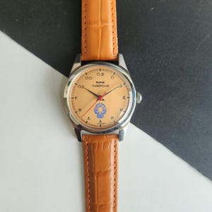 K&ouml;nnte beinhalten: Eine Armbanduhr mit hellbraunem Zifferblatt, silbernem Geh&auml;use und braunem Lederarmband. Das Zifferblatt hat schwarze Ziffern, rote und schwarze Zeiger und ein blaues Emblem. Das Armband ist hellbraun.