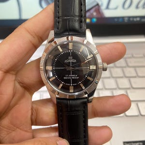 Puede incluir: Un reloj RW Roamer vintage con esfera negra y agujas e indicadores dorados. El reloj tiene una caja de acero inoxidable y una correa de cuero negro. La esfera del reloj incluye el texto "17 Jewels Shock Proof" y "Swiss Made".