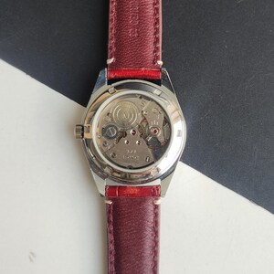 May include: A vintage wristwatch with a clear back, showcasing the intricate mechanical movement. The watch has a silver-toned case and a burgundy leather strap. The watch's internal gears and jewels are visible. The strap is stitched with white thread.