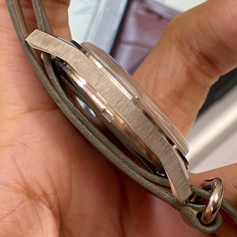 Puede incluir: Un reloj de pulsera plateado con una caja de metal texturizado y una correa de cuero gris. La esfera del reloj no es visible. La correa est&aacute; sujeta a la caja del reloj con un lazo de metal plateado.