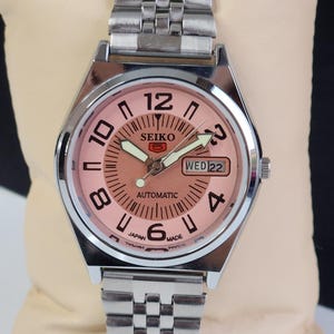 May include: A silver Seiko automatic wristwatch with a pink face, black numerals, and a day and date window. The watch has a silver metal band.