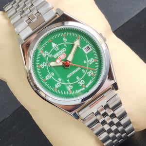 Könnte beinhalten: Eine silberne Seiko Automatik-Armbanduhr mit grünem Zifferblatt und silbernem Metallband. Das Zifferblatt der Uhr hat weiße Markierungen und rote Akzente. Das Zifferblatt der Uhr trägt außerdem den Text "SEIKO AUTOMATIC" und "JAPAN MADE".