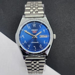 Könnte beinhalten: Eine silberfarbene Seiko 5 Automatik-Armbanduhr mit einem leuchtend blauen Zifferblatt. Die Uhr verfügt über arabische Ziffern, eine Tages-Datumsanzeige und ein Edelstahlarmband. Das Zifferblatt zeigt "MON 25".