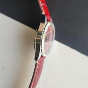 May include: A wristwatch with a red face and a silver-colored bezel. The watch has a red leather strap. The watch face displays the time with silver-colored hands and markers. The watch is angled to show the side profile.