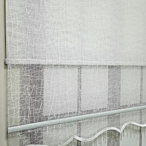 May include: White sheer window blind with a beaded fringe trim. The blind has a delicate, textured pattern that resembles a spiderweb.