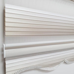 Op de afbeelding: Witte en beige gestreepte raambekleding met een gegolfde onderrand. De jaloezieën zijn gemaakt van een transparante stof en hebben een wit trekkoord.