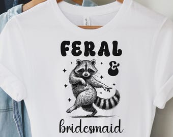 May include: White t-shirt with the text "FERAL & bridesmaid" and a raccoon graphic. The raccoon is standing and pointing. The word "WHITE" is in the top left corner.