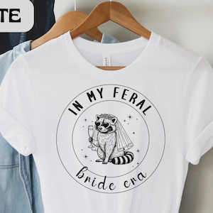 May include: White t-shirt with a black and white graphic of a raccoon wearing sunglasses and a veil, holding a champagne glass. The text "IN MY FERAL bride era" is in a circle around the raccoon. The word "WHITE" is in a white box in the upper left corner.