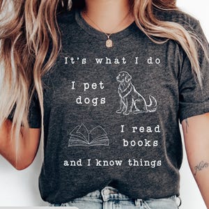 May include: Dark gray t-shirt with white text and illustrations. The text reads "It's what I do, I pet dogs, I read books, and I know things." A line drawing of a dog and an open book are also featured. The words "DARK GRAY" are at the bottom.