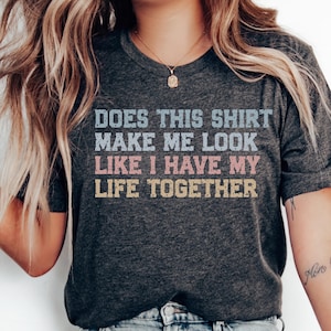 May include: A dark gray short-sleeved t-shirt featuring the text "DOES THIS SHIRT MAKE ME LOOK LIKE I HAVE MY LIFE TOGETHER" printed in a distressed, multi-colored font with light blue, pink, and yellow lettering across the chest area.