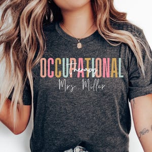 May include: Dark gray t-shirt with the words "OCCUPATIONAL Therapy Mrs. Miller" in colorful block letters. The shirt is a crew neck and made of a soft material. The text "DARK GRAY" is at the bottom of the image.