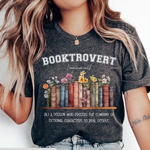 May include: Dark gray t-shirt with the word "BOOKTROVERT" in white, above a row of illustrated books and flowers. The definition of "booktrovert" is printed below. The shirt is dark gray.