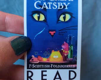 May include: A blue and white bookmark with a cat illustration. The text "The Great Gatsby" is at the top of the bookmark. The text "F. SCOTTISH-FOLDZGERALD" is at the bottom of the bookmark. The word "READ" is in large black letters at the bottom of the bookmark.