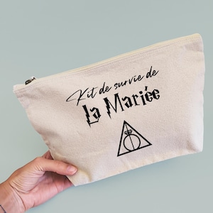 Può includere: Pochette con cerniera in tela beige con testo nero che recita "Kit de survie de La Mariée" e un grafico di un triangolo con un anello all'interno. La pochette è tenuta da una persona.