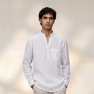 May include: A white linen shirt with a mandarin collar and a chest pocket, paired with light beige linen pants. The outfit is completed with blue sandals. The clothing is lightweight and suitable for warm weather.