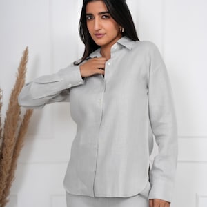 Puede incluir: Una camisa de lino gris claro y pantalones a juego. La camisa abotonada de manga larga está parcialmente desabrochada. El conjunto es de color neutro y parece estar hecho de una tela ligera. Adecuado para ropa casual o de negocios.