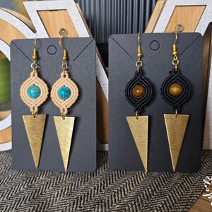 Peut inclure: Deux paires de boucles d'oreilles en macramé sur des supports noirs. Une paire présente un cordon beige avec des perles turquoise et des pendentifs triangulaires dorés. L'autre paire a un cordon noir avec des perles brunes et des pendentifs dorés.