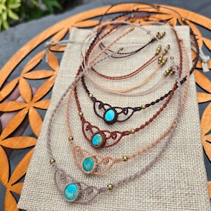 Puede incluir: Una colección de collares de macramé hechos a mano con piedras turquesas. Los collares presentan intrincados nudos en tonos marrones y beige, acentuados con pequeñas cuentas doradas. Se exhiben sobre una tela tipo arpillera.
