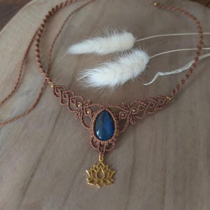 Peut inclure: Un collier ras de cou en macramé marron avec un pendentif en pierre bleue et un charme en forme de lotus doré.