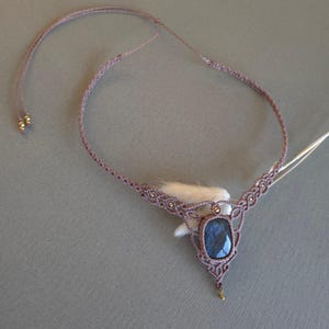 Puede incluir: Un delicado collar de macramé de color malva con un gran colgante de labradorita azul oscuro. El collar está adornado con pequeñas cuentas doradas.
