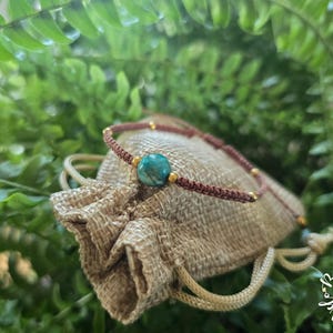 May include: A brown cord bracelet featuring a central round turquoise-colored stone bead, accented with small gold-toned metallic beads along the band, resting on a small, textured beige fabric drawstring pouch.