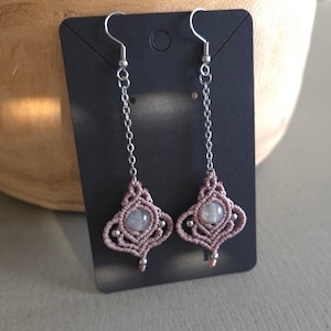 Peut inclure: Une paire de longues boucles d'oreilles en chaîne argentée avec un motif en macramé rose et une perle de pierre de lune blanche au bas.