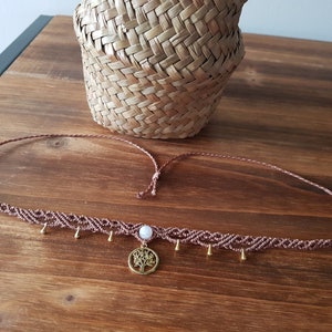 Könnte beinhalten: Eine braune Makramee-Halskette mit einem goldenen Lebensbaum-Anhänger und einer weißen Perle. Die Halskette hat kleine goldene Perlen, die daran hängen.