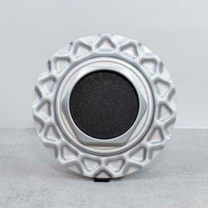 Może przedstawiać: Srebrny, okrągły przedmiot z czarnym, teksturowanym środkiem. Zewnętrzna krawędź ma powtarzający się geometryczny wzór. Obiekt jest prawdopodobnie elementem dekoracyjnym lub funkcjonalnym, być może do pojazdu lub maszyny.