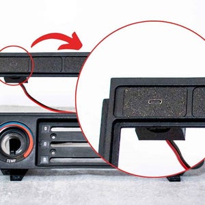 以下が含まれることがあります： USB-Cポート、赤と黒のワイヤー、温度調節ダイヤルを備えた黒い車のダッシュボードコンポーネント。 コンポーネントはテクスチャード加工された表面と長方形の形状をしています。 ダイヤルは青と赤です。