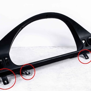 May include: A black car dashboard instrument cluster bezel with an arched top and a rectangular bottom. It has several openings for gauges and controls, and mounting brackets are visible.