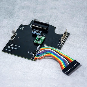 Könnte beinhalten: Schwarze Leiterplatte mit verschiedenen elektronischen Bauteilen, darunter ein kleines grünes Modul, ein Kondensator und mehrere Stiftleisten. Ein farbenfrohes Flachbandkabel mit einem schwarzen Stecker ist angeschlossen. Die Platine trägt den Text "MOD-ING".