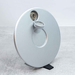 Könnte beinhalten: Ein runder Tankdeckel aus Silber mit einem Schlüsselloch und einer kleinen abnehmbaren silbernen Kappe. Der Deckel liegt auf einer grauen Oberfläche.