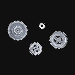 May include: A collection of small, transparent plastic gears of different sizes, set against a black backdrop. The gears feature toothed rims and central apertures, indicating they are parts of a mechanism or model.
