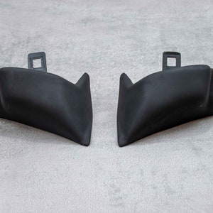 Könnte beinhalten: Zwei schwarze Kunststoff-Stoßfängerecken. Die Teile sind so konzipiert, dass sie an die Ecken eines Autostoßfängers passen und oben ein kleines Loch haben.