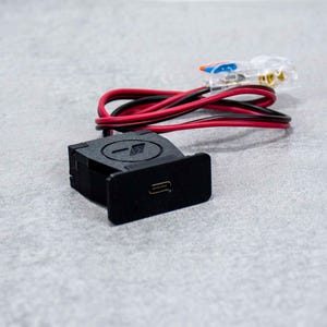 Könnte beinhalten: Ein schwarzes elektrisches Bauteil mit einem rot-schwarzen Kabel. Das Bauteil hat einen kleinen rechteckigen Knopf mit einem Symbol darauf.