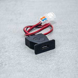 Op de afbeelding: Een zwarte USB-adapter met een rood en zwart snoer eraan. De adapter heeft een klein rond logo met de letter "T" erin. Het snoer heeft een RJ-11-connector aan het andere uiteinde.