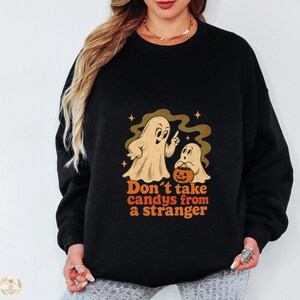 Könnte beinhalten: Ein schwarzes Langarm-Sweatshirt mit einer Grafik von zwei Geistern, einem größeren und einem kleineren, der einen Kürbiseimer hält. Der Text unter den Geistern lautet "Don't take candys from a stranger" in orangefarbenen Buchstaben mit kleinen Sternen.