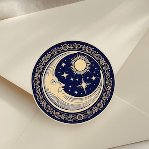 Könnte beinhalten: Ein runder Aufkleber mit dunkelblauem Hintergrund, der einen cremefarbenen Halbmond mit Gesicht, eine Sonne und Sterne zeigt. Das Design ist von einem verzierten, kreisförmigen goldfarbenen Rand mit komplexen Schnörkeln und Blumenmustern umgeben.