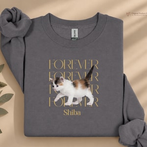 Könnte beinhalten: Ein anthrazitfarbenes Sweatshirt mit Rundhalsausschnitt und einer Grafik eines laufenden Glückskätzchens. Der Text "FOREVER" ist viermal in einer goldenen Serifenschrift hinter dem Kätzchen wiederholt, darunter steht das Wort "Shiba" in Gold.